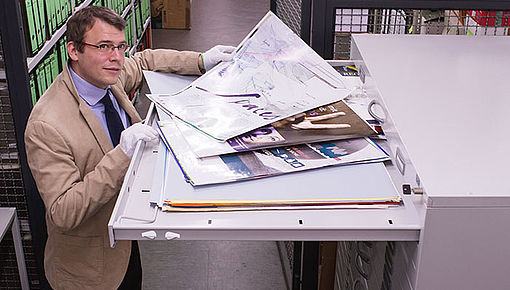 Universitätsarchivar Dr. Becker öffnet einen Archivschrank, in dem eine Reihe von Universitätsplakaten zu sehen ist. Foto: Referat Kommunikation, M. Weich