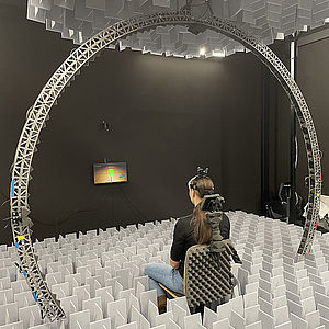 Figure 1: measurement setup for the individual HRTF: subjects take a seat at the center of the arc and are exposed to sound from 24 different loudspeakers in all hearing-relevant frequencies while the arch rotates around the subjects. © Sarah Roßkopf