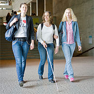 Eine sehbehinderte Studentin mit Blindenstock wird von zwei sehenden Studentinnen durch das Zentrale Hörsaalgebäude begleitet