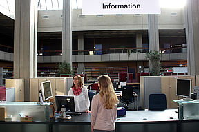 Das Bild zeigt den Informationsschalter von der Uni-Bibliothek. Man sieht eine Mitarbeiterin, die einer Studentin etwas erklärt.