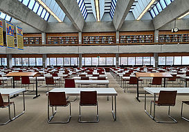 Das Bild zeigt einen Lesesaal in der Uni-Bibliothek. Man sieht einen großen, hellen Raum. Darin stehen viele Tische und Stühle. Im Hintergrund sieht man Studentinnen und Studenten, die arbeiten. Man sieht auch einige Bücherregale.