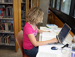 Das Bild zeigt einen Arbeitsplatz in der Uni-Bibliothek. Eine Studentin sitzt an einem Tisch und liest ein Buch. Auf dem Tisch steht ein Laptop. Man sieht auf dem Tisch auch eine Flasche Wasser und mehrere Bücher.