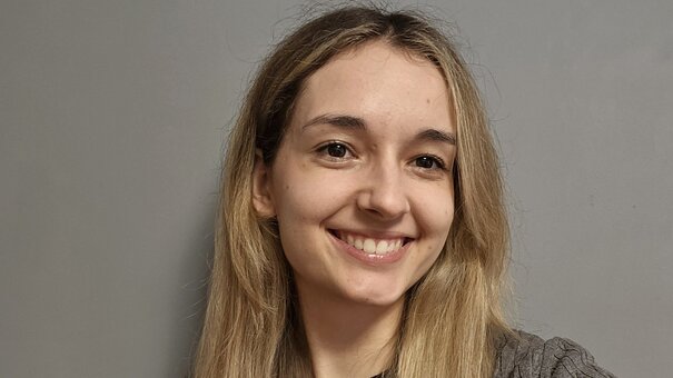 Ein Porträt einer blonden jungen Frau mit langen Haaren vor einem grauen Hintergrund.