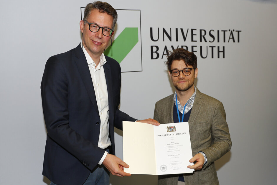 Zwei Männer in formeller Kleidung schauen in Richtung Betrachter und halten gemeinsam eine geöffente Urkunden-Mappe. Auf dem Dokument in der Mappe ist das Bayerische Staatswappen zu erkennen. (öffnet Vergrößerung des Bildes)