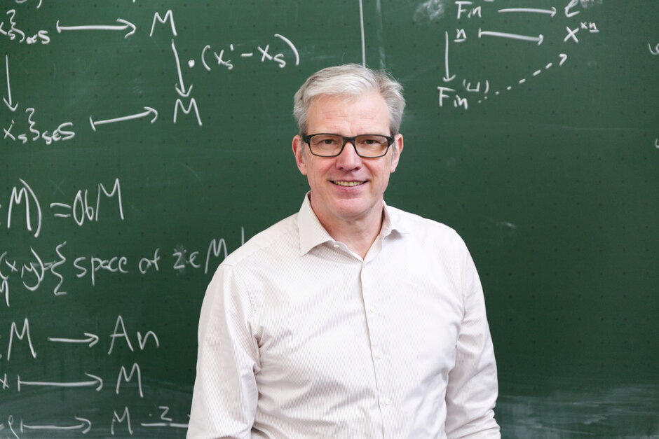 Ein Mann mit grauen, kurzen Haaren, Brille mit schwarzem Rand und einem weißen Hemd steht vor einer grünen Wandtafel auf der mit weißer Kreise mathematische Formeln notiert wurden.