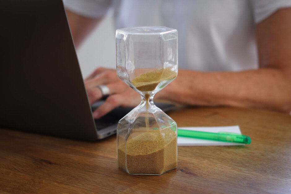 Sanduhr im Vordergrund, im Hintergrund eine Person, die etwas auf einem Laptop schreibt