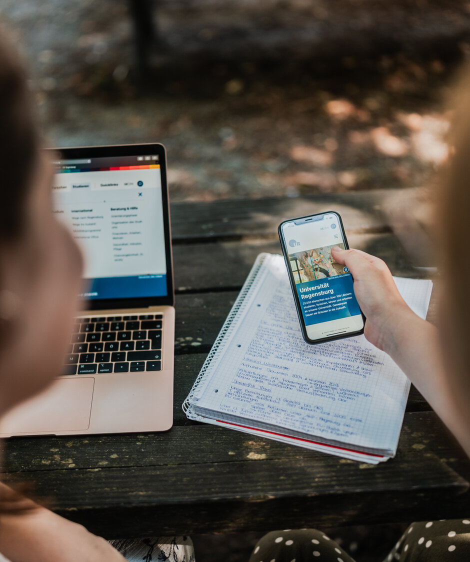 Zwei Studierende blicken gemeinsam auf das Display eines Smartphones. Im Hintergrund sind ein Laptop und ein beschriebener Notizblock zu sehen.