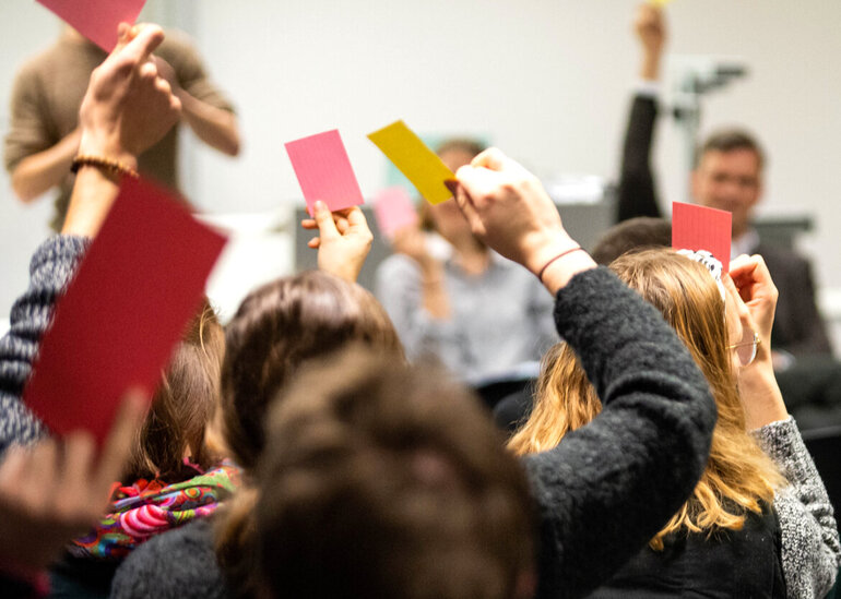 In einem Seminarraum halten Menschen farbige Karten in gelb oder rot zur Abstimmung in die Höhe. 