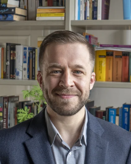 Porträtfoto eines Mannes. Im Hintergrund ist ein Bücherregal zu sehen. (öffnet Vergrößerung des Bildes)