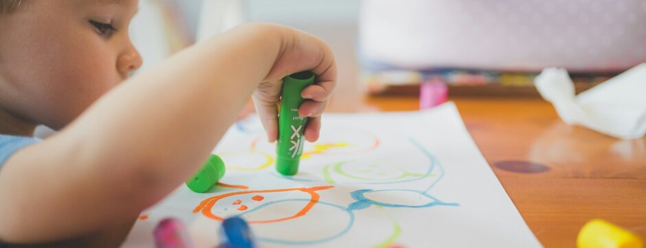Kleinkind sitzt am Tisch und malt, bunte STifte liegen verteilt auf dem Tisch
