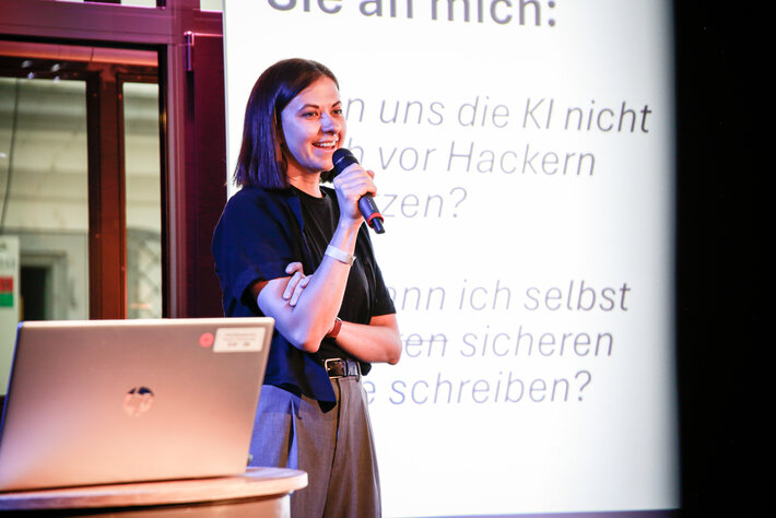 Person steht auf einer Bühne vor einer großen Projektionsfläche, hält ein Mikrofon und hat den anderen Arm locker verschränkt. Neben der Person steht ein Laptop auf einem kleinen Tisch beziehungsweise Fass. Auf der Leinwand im Hintergrund ist unscharfer deutscher Text zu sehen, unter anderem eine Frage zu „KI“ und „Hackern“. Die Szene ist in farbiges Bühnenlicht getaucht, mit dunklen Vorhängen und Fenstern im Hintergrund. (öffnet Vergrößerung des Bildes)