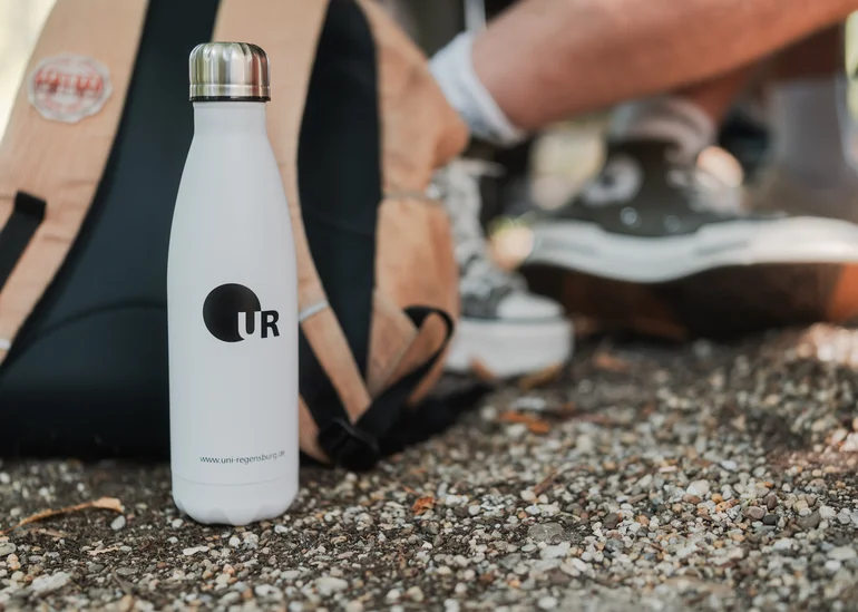 Das Bild zeigt eine weiße Trinkflasche mit UR-Logo, die vor einem Rucksack steht, der neben den Füßen einer am Boden sitzenden Person liegt..