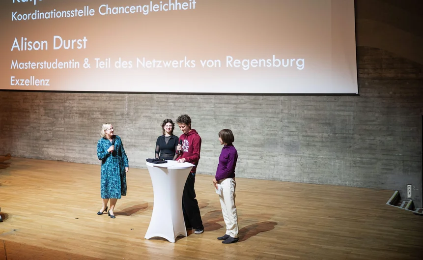 Vier Personen stehen auf einer Bühne um ein weißes Rednerpult. Auf der großen Leinwand im Hintergrund werden Informationen zur Koordinationsstelle Chancengleichheit angezeigt.  (öffnet Vergrößerung des Bildes)