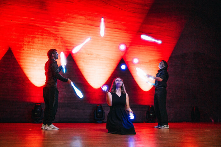 Drei junge Menschen jonglieren auf der Audimax-Bühne mit leuchtenden Kegeln und Bällen. Der Hintergrund ist rot beleuchtet. (öffnet Vergrößerung des Bildes)