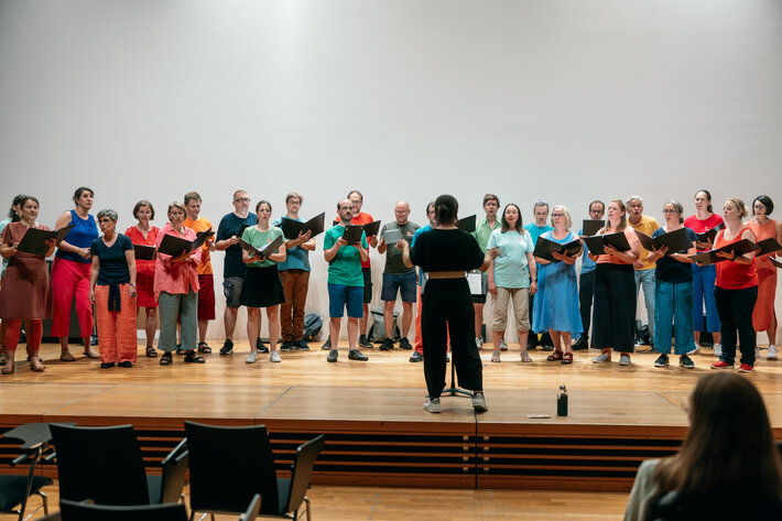 Bunt gekleidete Menschen stehen auf der Bühne und singen. Sie werden dirigiert von einer Frau in schwarzer Kleidung. (öffnet Vergrößerung des Bildes)
