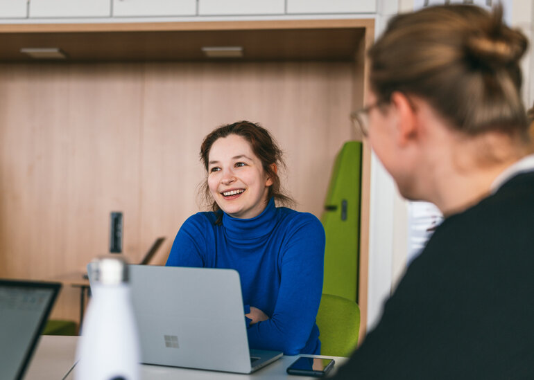 Eine Studentin sitzt lächelnd und konentriert zusammen mit anderen Studierenden an einem Seminartisch