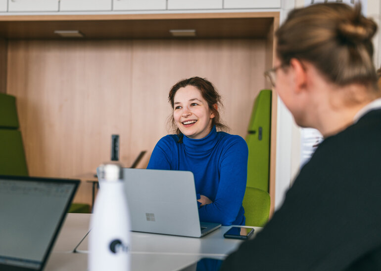 Eine Studentin sitzt lächelnd und konentriert zusammen mit anderen Studierenden an einem Seminartisch