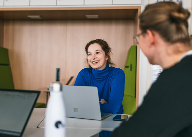 Eine Studentin sitzt lächelnd und konentriert zusammen mit anderen Studierenden an einem Seminartisch