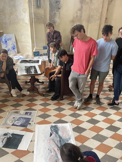 Mehrere Personen befinden sich in einem Raum mit gemustertem Boden und abgenutzten Wänden. Sie stehen oder sitzen um einen Tisch und betrachten verschiedene Kunstwerke, die auf dem Tisch und dem Boden verteilt sind. Die Werke zeigen unterschiedliche Stile und Motive, und die Personen wirken in Gespräche vertieft. (öffnet Vergrößerung des Bildes)