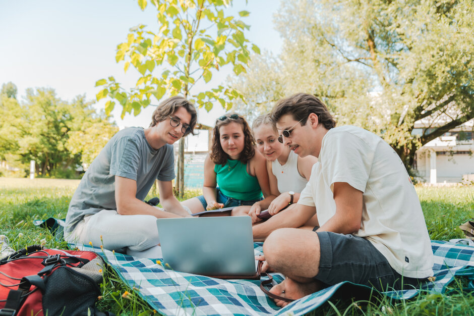 Vier Studierende sitzen auf einer Picknickdecke auf einer Wiese am Campus der Universität Regensburg und schauen gemeinsam in einen Laptop. (externer Link, öffnet neues Fenster)