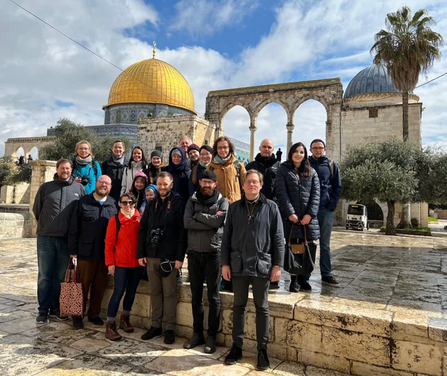 Rund 20 Personen in warmen Jacken stehen im Sonnenschein auf einem Platz, der mit historischen Gebäuden umgeben ist. Im Hintergrund sind Palmen und Olivenbäume zu erkennen sowie eine gold-gläzende Kuppel. (öffnet Vergrößerung des Bildes)