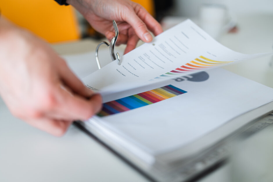 Zwei Hände ordnen ein farbig bedrucktes DINA 4-Papier in einen Pappordner ein.