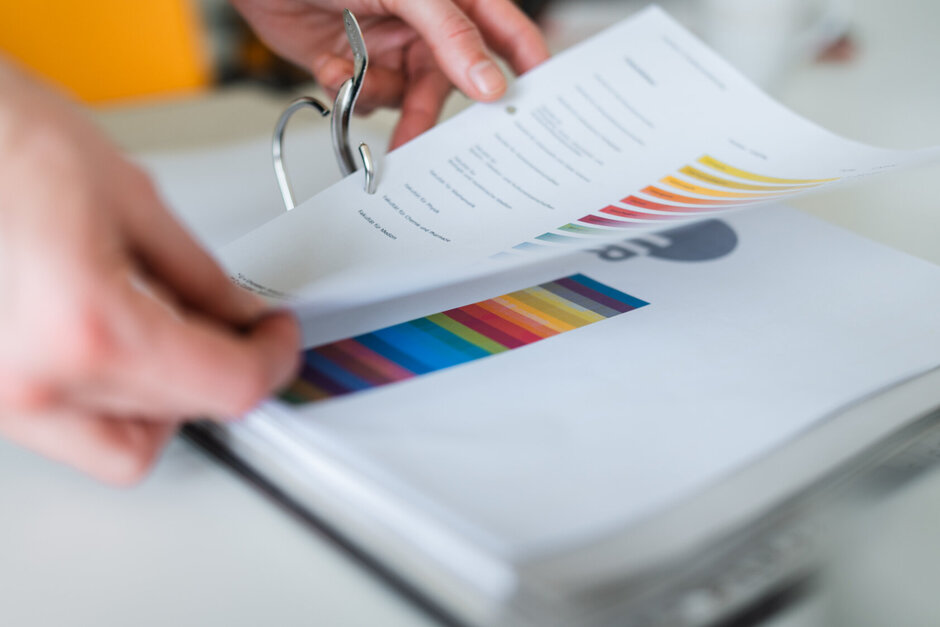 Zwei Hände ordnen ein farbig bedrucktes DINA 4-Papier in einen Pappordner ein.