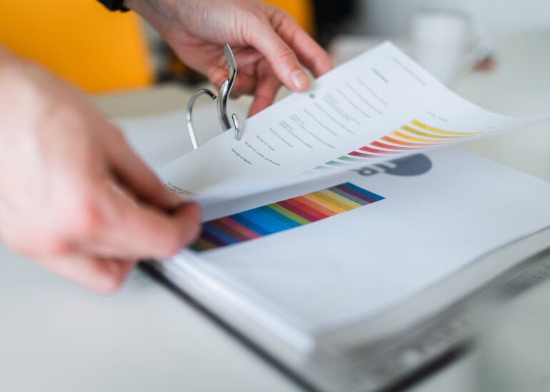 Zwei Hände ordnen ein farbig bedrucktes DINA 4-Papier in einen Pappordner ein.