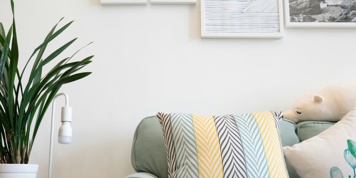 Zu sehen ist eine hellgraue Couch mit bunten Kissen. Links von der Couch steht ein kleines Tischchen mit einer Grünpflanze in einem weißen Übertopf sowie einer weißen Lampfe. Über der Couch hängen Bilder in weißer und grauer Farbe.