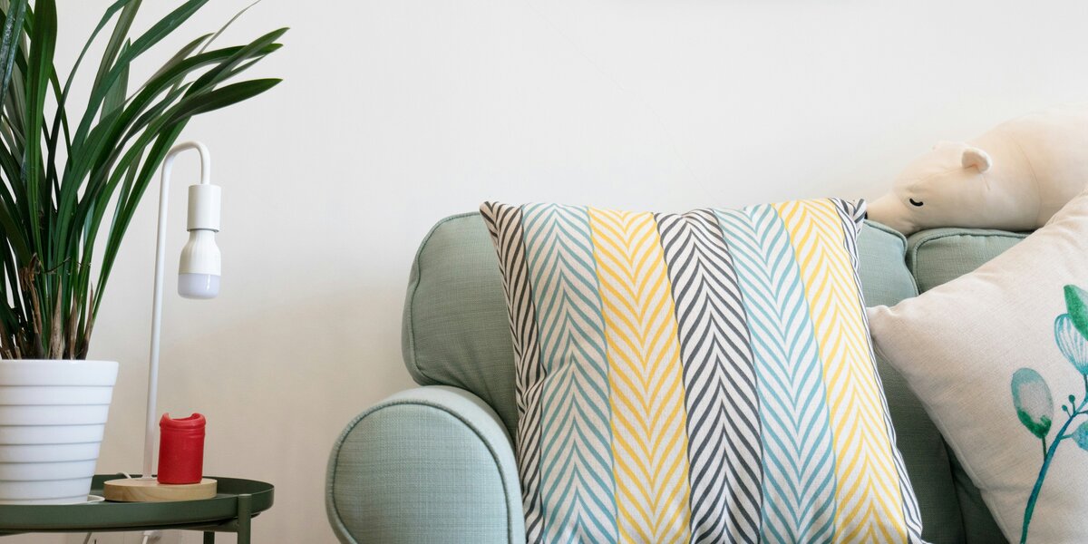 Zu sehen ist eine hellgraue Couch mit bunten Kissen. Links von der Couch steht ein kleines Tischchen mit einer Grünpflanze in einem weißen Übertopf sowie einer weißen Lampfe. Über der Couch hängen Bilder in weißer und grauer Farbe.