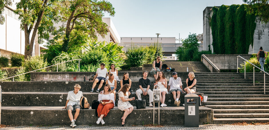 Eine große Gruppe Studierender sitzt im Sommer vor dem Gebäudeteil Philosophie/Theologie der Universität Regensburg.