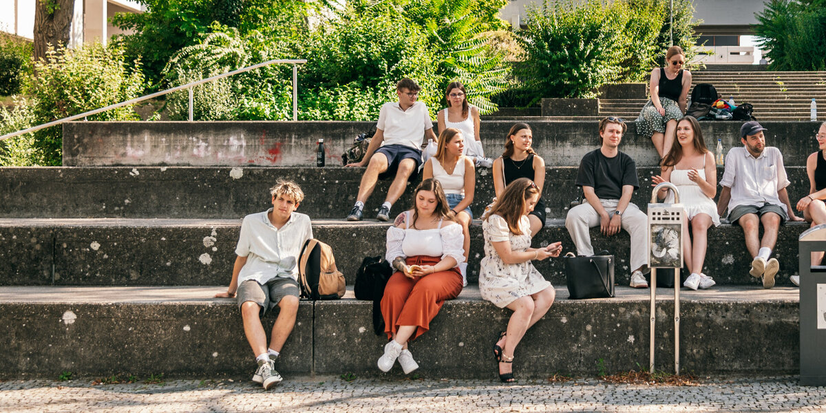 Eine große Gruppe Studierender sitzt im Sommer vor dem Gebäudeteil Philosophie/Theologie der Universität Regensburg.