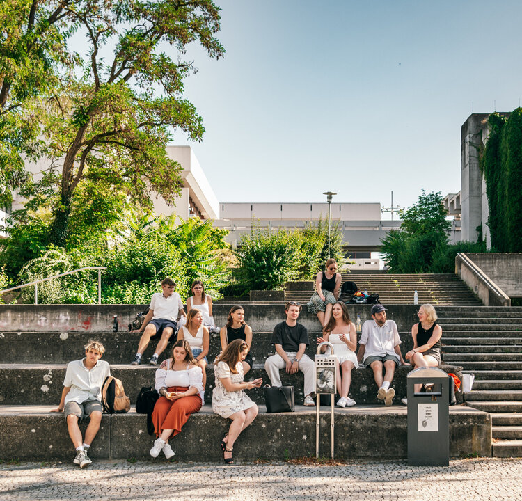 Eine große Gruppe Studierender sitzt im Sommer vor dem Gebäudeteil Philosophie/Theologie der Universität Regensburg.