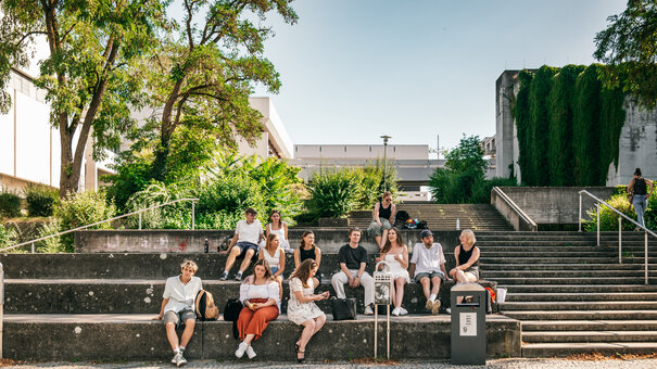 Eine große Gruppe Studierender sitzt im Sommer vor dem Gebäudeteil Philosophie/Theologie der Universität Regensburg.