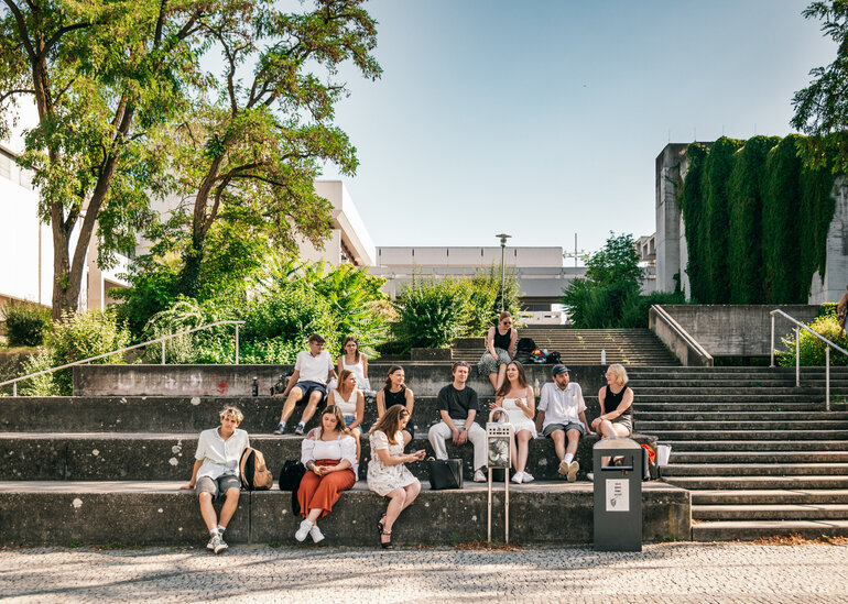 Eine große Gruppe Studierender sitzt im Sommer vor dem Gebäudeteil Philosophie/Theologie der Universität Regensburg.