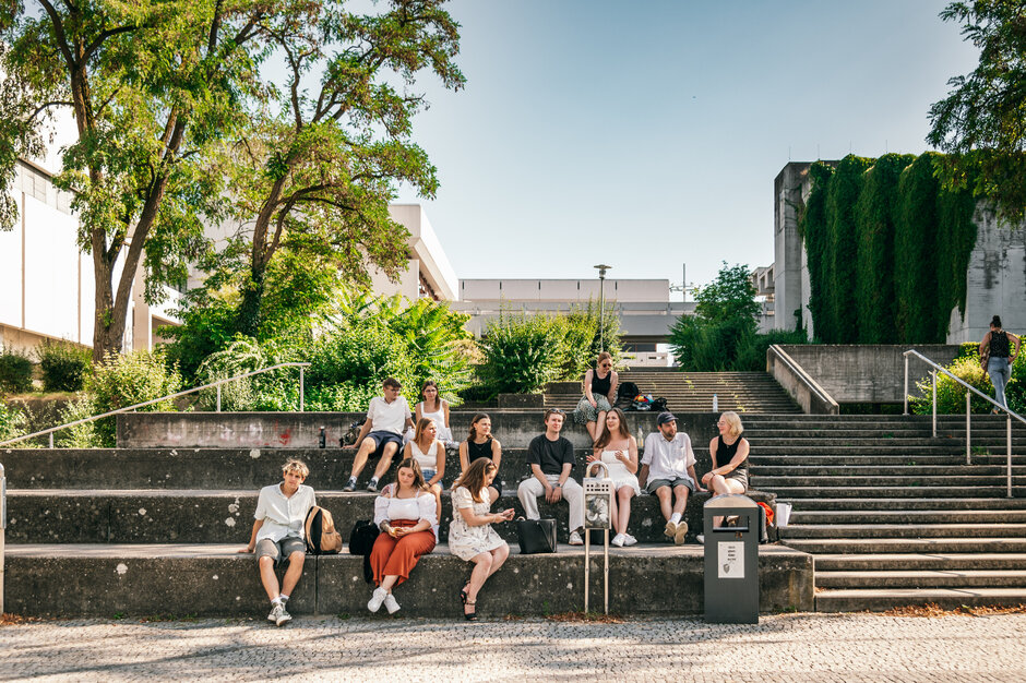 Eine große Gruppe Studierender sitzt im Sommer vor dem Gebäudeteil Philosophie/Theologie der Universität Regensburg.