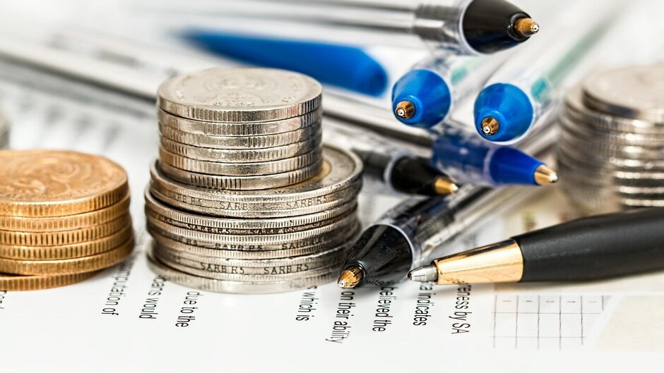 Man sieht Münzen auf einem Stück Papier gestapelt sowie mehrere Kugelschreiber