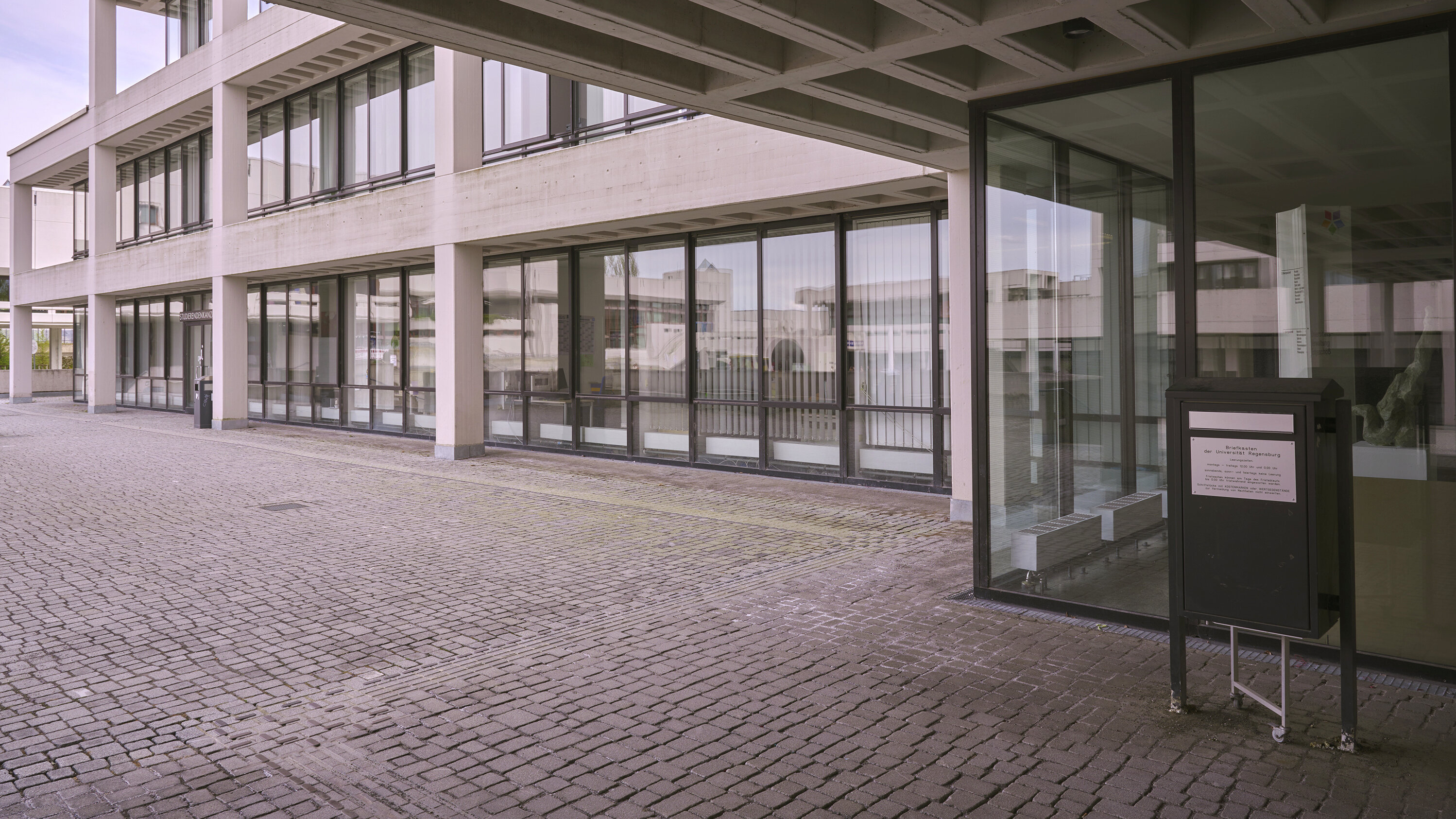 Eingangsbereich eines Gebäudekomplexes in Sichtbetonbauweise; vor einer Glasfront ist ein großformatiger Briefkasten zu sehen. (öffnet Vergrößerung des Bildes)