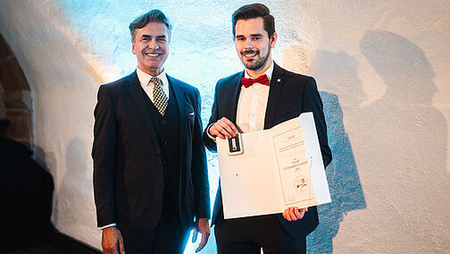 Zwei Männer stehen vor einer weißen Wand. Der links Stehende trägt einen dunklen Dreiteiler und Krawatte, der rechts Stehende trägt einen dunklen Anzug mit roter Fliege. Er hält eine aufgeschlagene Urkundenmappe in Händen und zeigt sie den Betrachtern. 