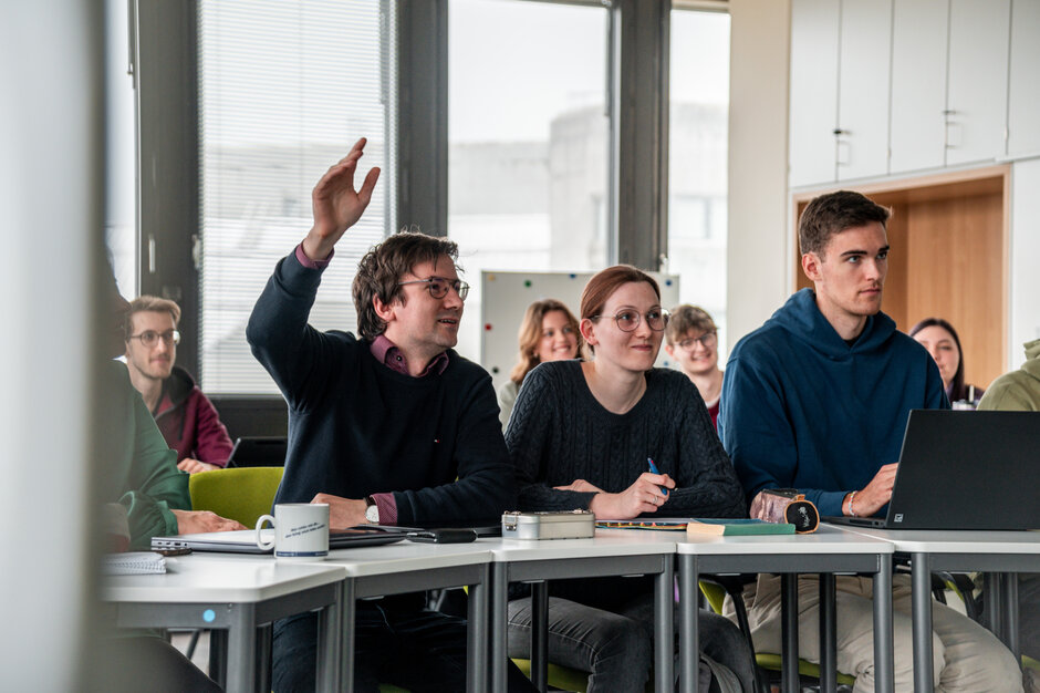 Studierende in einem Seminarraum. Ein Student meldet sich mit erhobener Hand.