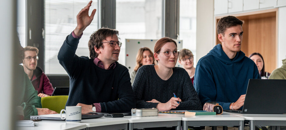 Studierende in einem Seminarraum. Ein Student meldet sich mit erhobener Hand.