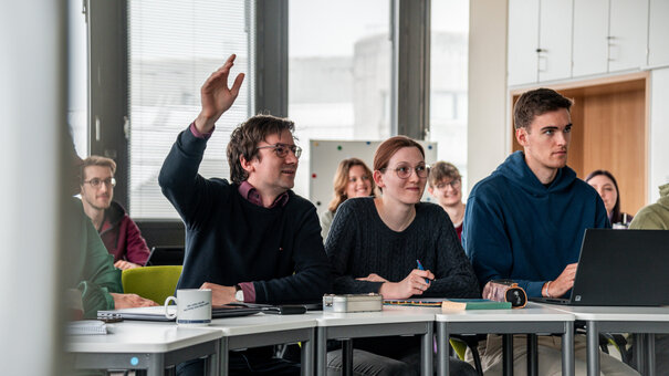 Studierende in einem Seminarraum. Ein Student meldet sich mit erhobener Hand.