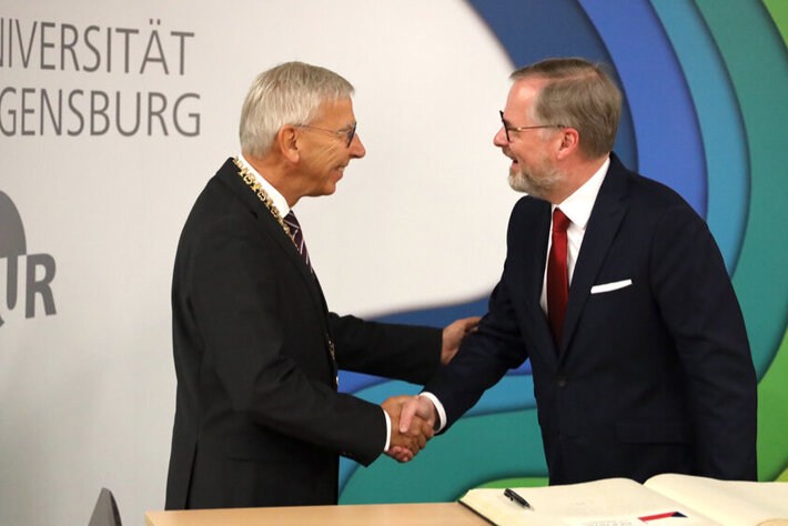 Zwei Männer in formeller Kleidung schütteln sich die Hand. Im Hintergrund ein Aufsteller mit dem Aufdruck "Universität Regensburg" und dem Logo der Universität. (öffnet Vergrößerung des Bildes)