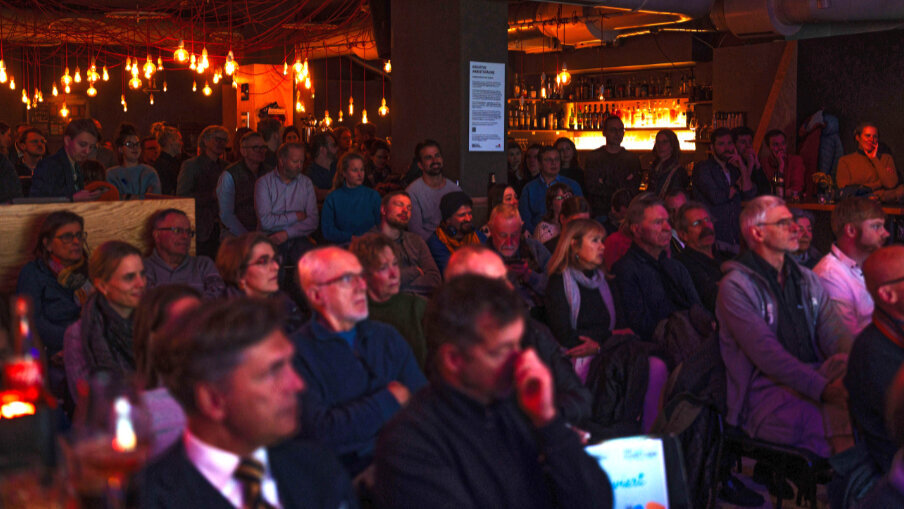 Ein stimmungsvoll beleuchteter, voll besetzter Veranstaltungsraum. Das Publikum schaut gespannt Richtung Bühne.
