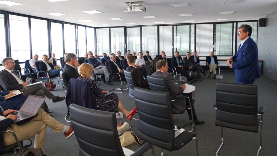 Ein Konferenzraum mit großen Fenstern, in dem zahlreiche Personen in einem Halbkreis sitzen und einem Vortragenden im blauen Anzug zuhören; moderne Bürostühle, einige Zuhörer mit Laptops oder Notizblöcken. (öffnet Vergrößerung des Bildes)