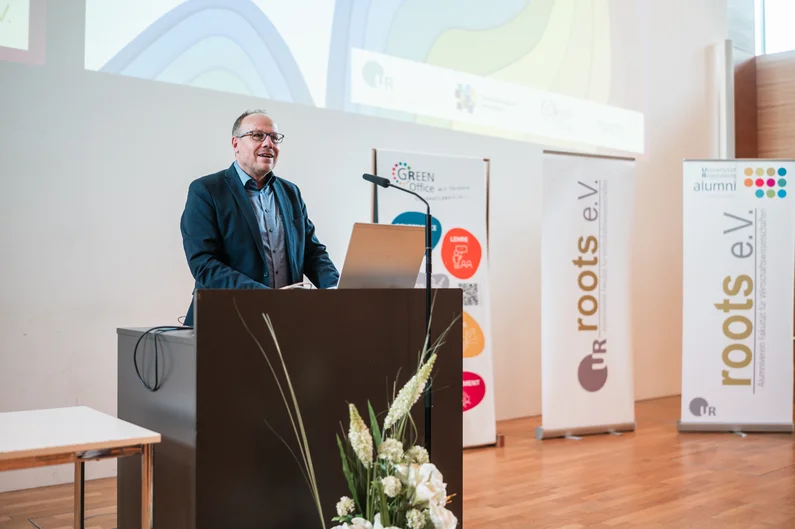 Ein Mann mit Brille und im Anzug steht an einem Rednerpult und spricht. Im Hintergrund sind drei Aufsteller zu sehen. Auf zwei Aufstellern steht "roots e.V.", auf dem dritten steht "Green Office". (öffnet Vergrößerung des Bildes)