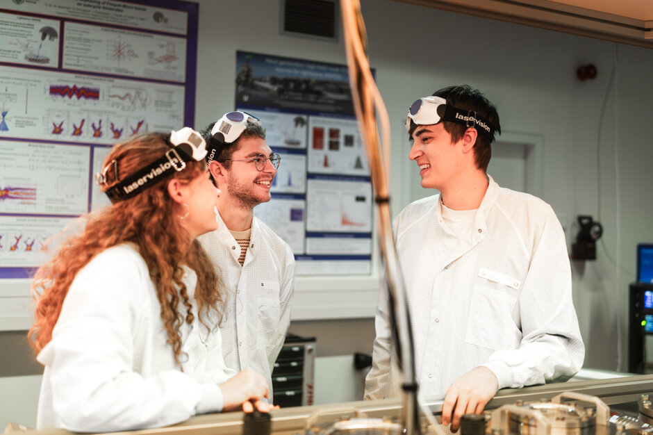 Drei Nachwuchswissenschaflter*innen mit weißen Laborkitteln und auf die Stirn geschobenen Schutzbrillen stehen in einem Labor und unterhalten sich lächelnd.