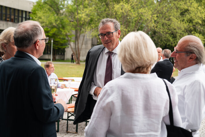 Ein Mann, der seine Anzugjacke leger über seinen Rücken gelegt hat, im Gespräch mit einer Gruppe von vier weiteren Personen. (öffnet Vergrößerung des Bildes)