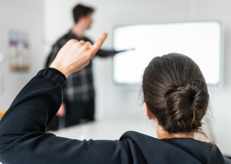 Eine Person meldet sich während ein Mann im Hintergrund etwas an einem Whiteboard zeigt.
