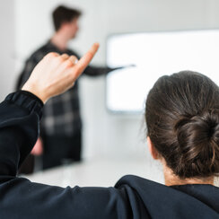 Eine Person meldet sich während ein Mann im Hintergrund etwas an einem Whiteboard zeigt.